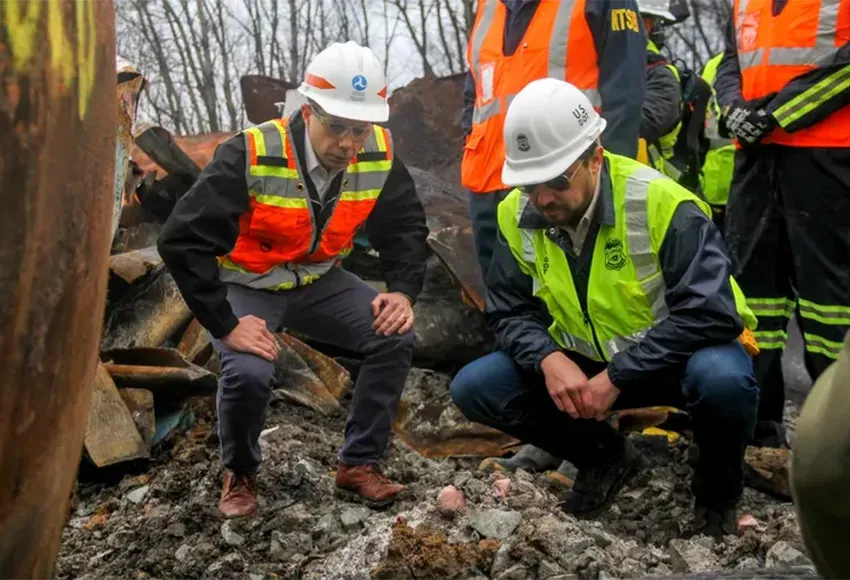 Transportation Secretary Pete Buttigieg (l) and Tristan Brown, deputy administrator of the Pipeline and Hazardous Materials Safety Administration, crouch down to look at part of a burned traincar on Feb. 23, 2023, in East Palestine, Ohio, at the site of a Norfolk Southern train derailment – Photo by Allie Vugrincic / The Vindicator via AP